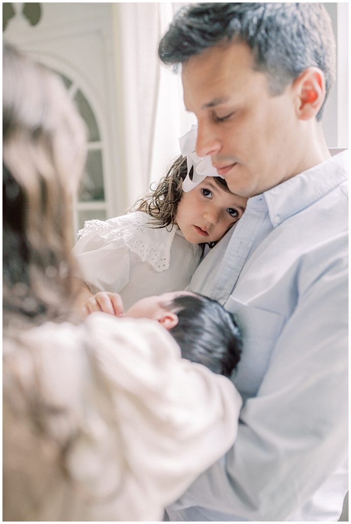 A Mother And Father Hold Their Newborn Daughter While Their Toddler Daughter Looks At The Camera.