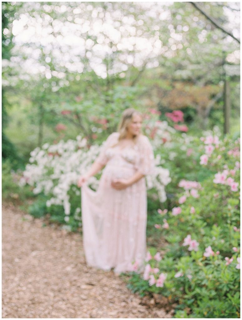 An Out-Of-Focus Image Of A Pregnant Mother In A Pink Dress Holding Her Dress And Cradling Her Belly In A Garden.