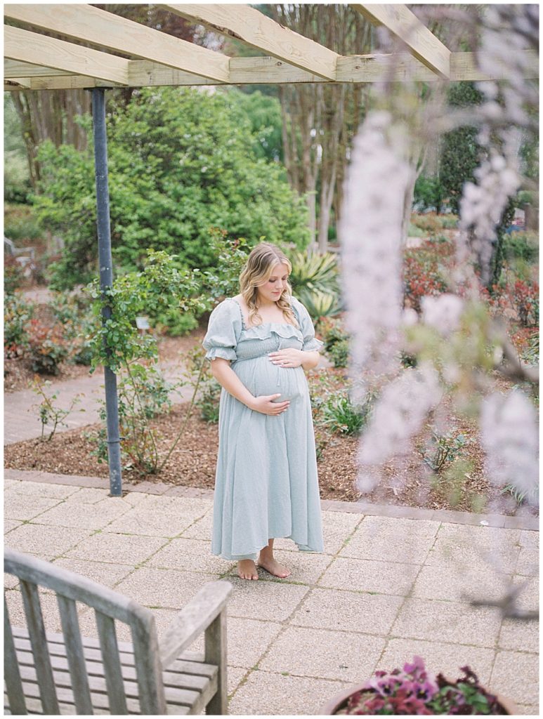 Blonde Pregnant Woman Stands Under A Wooden Pergola With Wisteria With One Hand Below Her Belly And One Hand Underneath.