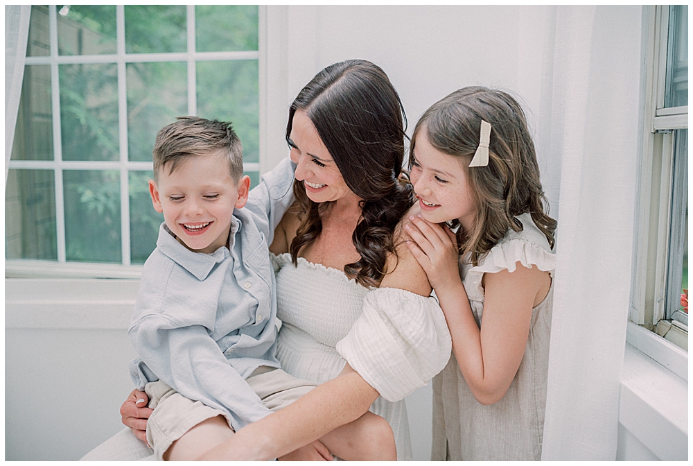 A Mother Sits On A Stool With Her Son On Her Lap And Her Daughter Standing Behind Her, Smiling At Them