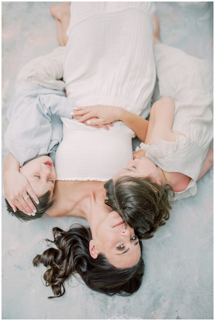 A Mother Lays Down On The Floor With Her Son And Daughter And Looks Directly At The Camera