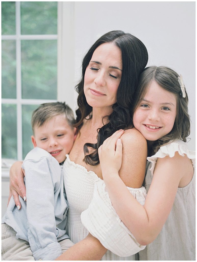 A Mother Sits On The Chair Holding Her Son While Her Daughter Stands Close, Higging Her