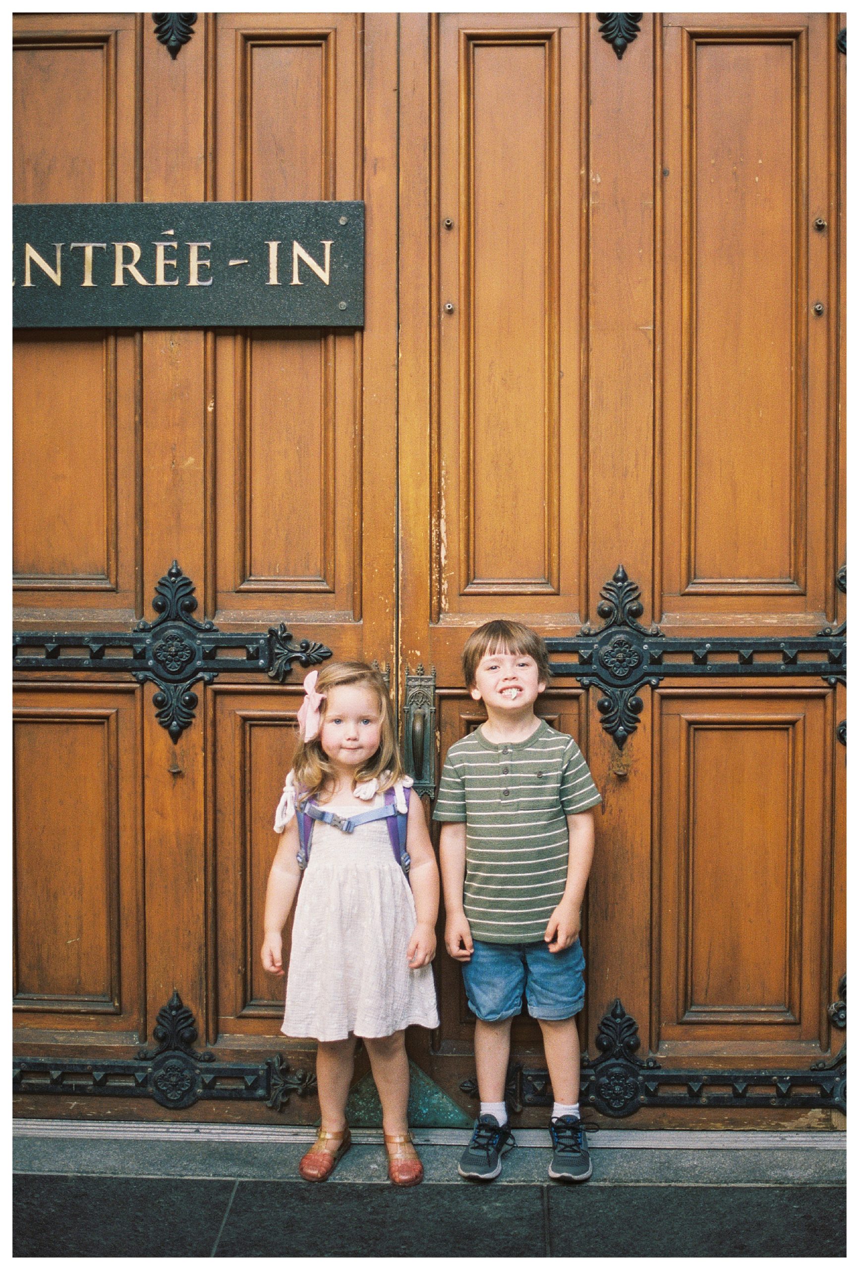 Two Toddlers Stand In Front Of Large Wooden Door At Notre Dame Cathedral In Old Montreal.