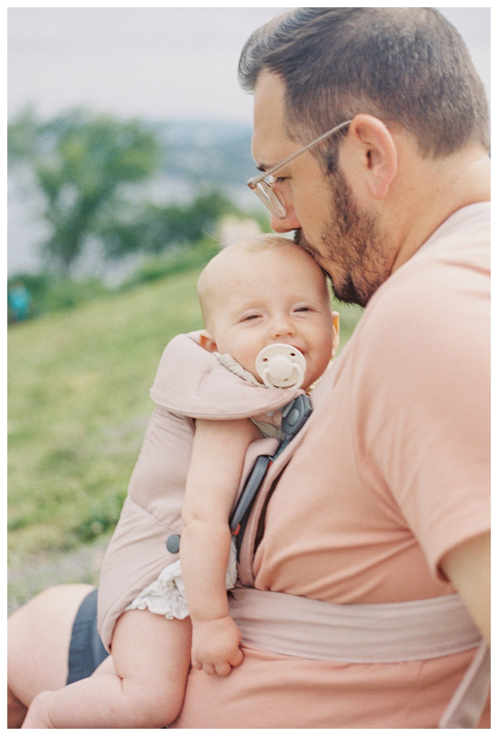 Father Lean Into His Infant Daughter Riding In A Carrier And Kisses Her Head As She Smiles.