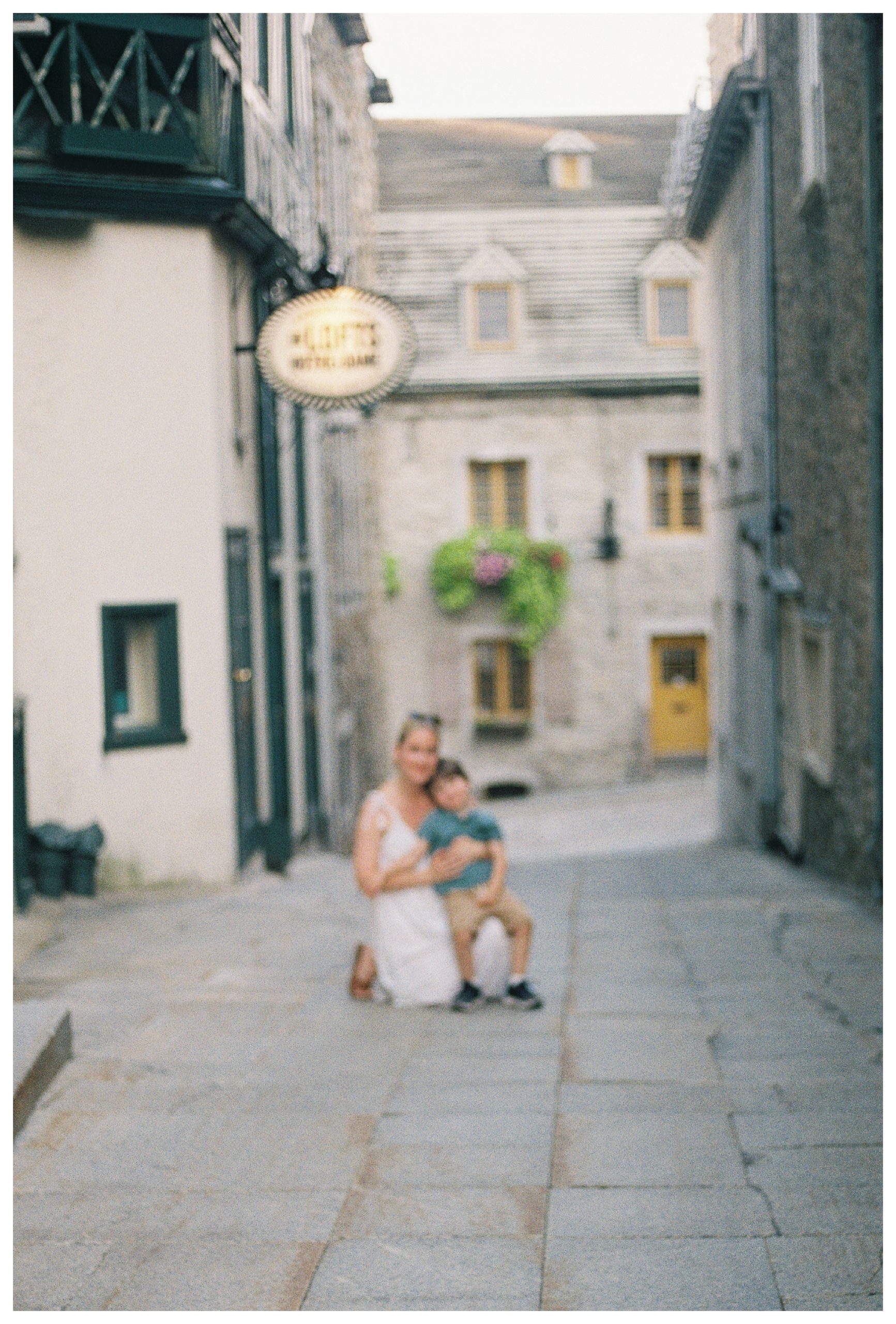 Blurry Image Of Mother In White Dress Kneeling Down On Quebec City Street While Her Toddler Son Sits In Her Lap.