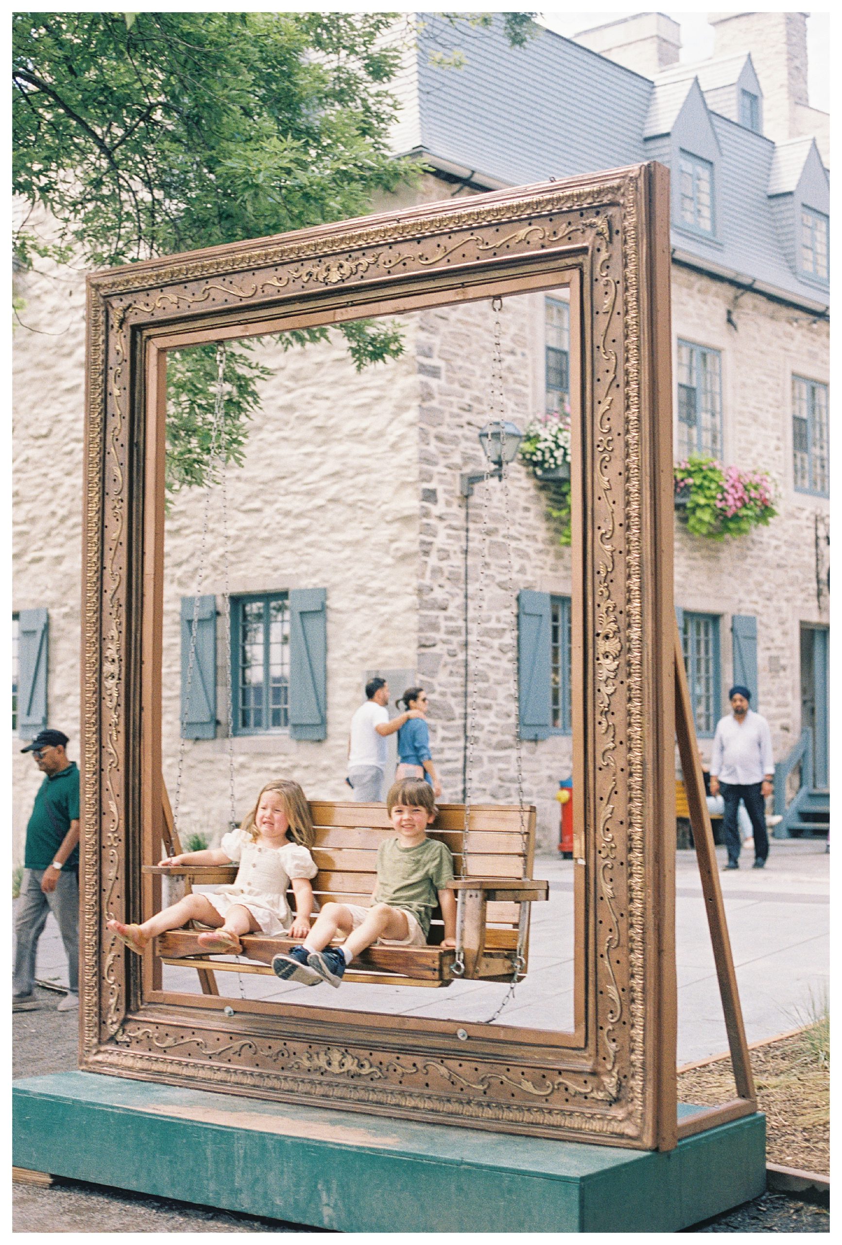 Two Toddlers Sit On A Swinging Bench, One Smiling, One Crying, In Quebec City