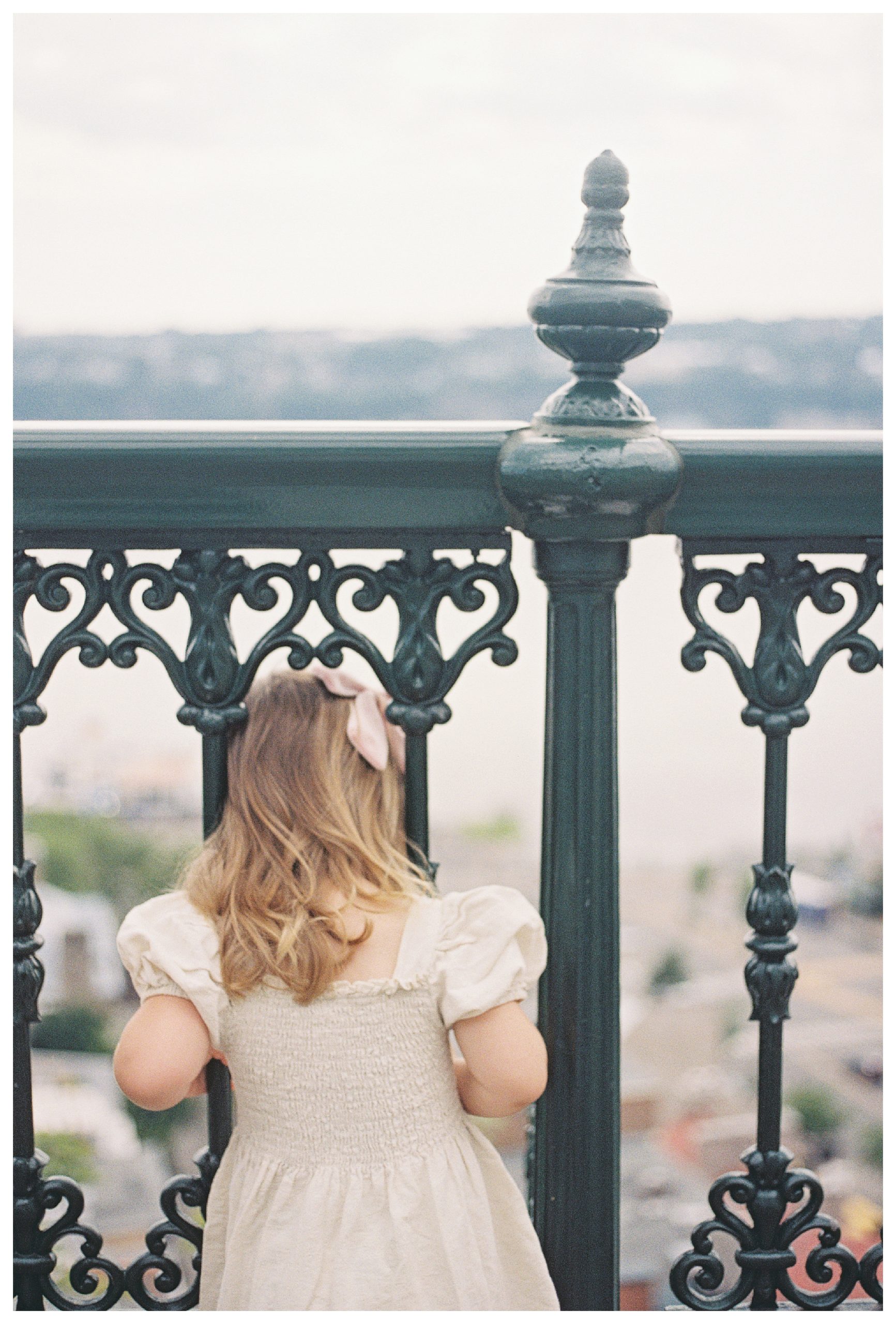 Little Girl With Brown Curly Hair Peaks Through Iron Railings In Quebec City