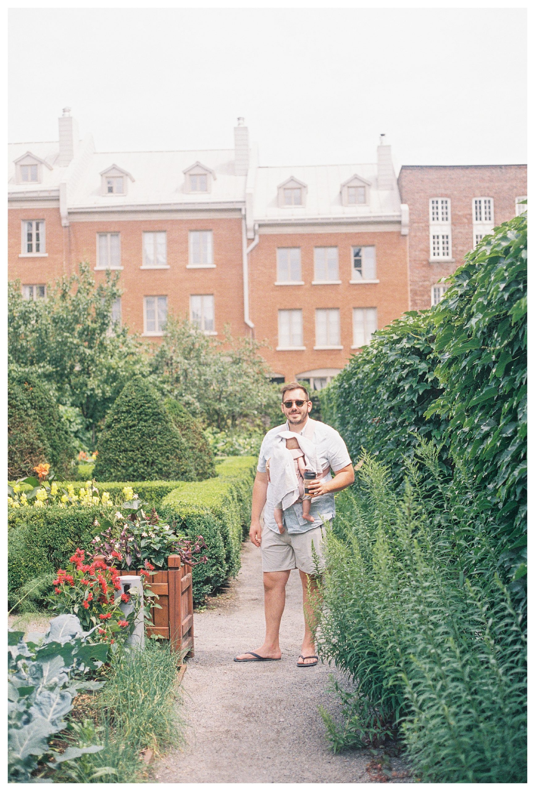 Father Wearing Sunglasses And Baby In Carrier Holds Coffee And Smiles In Garden In Montreal