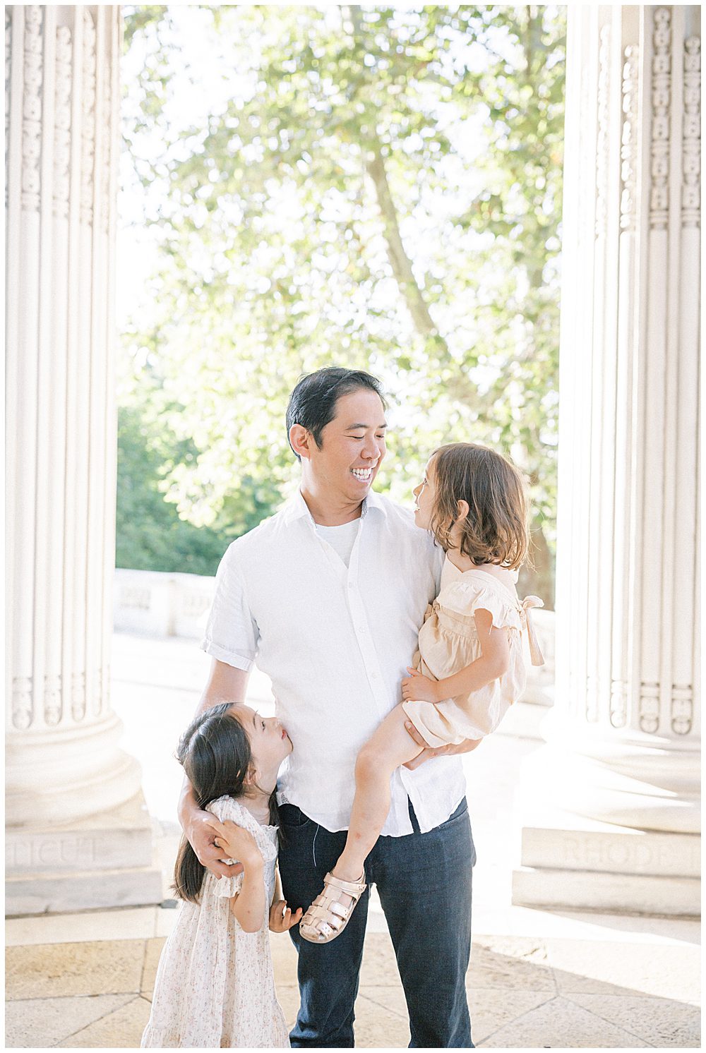 Father Holds One Daughter And Smiles At Her While Other Daughter Stands Next To Him, Looking Up At Him.