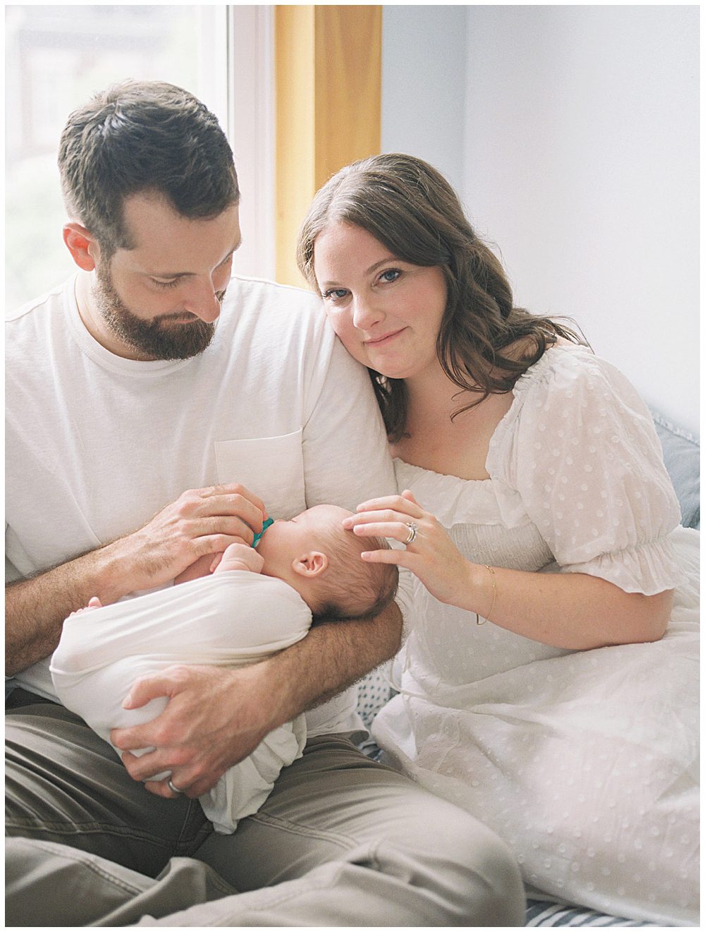 Mom Looks At The Camera With A Soft Smiles And Touches Her Baby's Head While Her Husband Holds Baby.