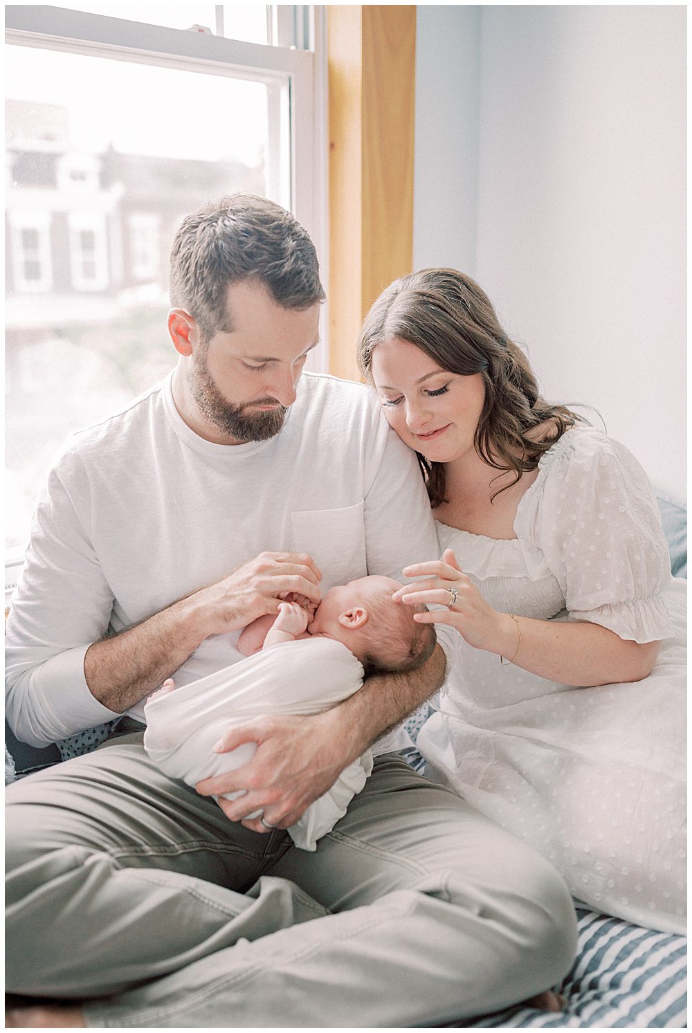 New Mom Gently Traces Her Newborn's Hair While Leaning Against Her Husband As They Sit On Their Bed