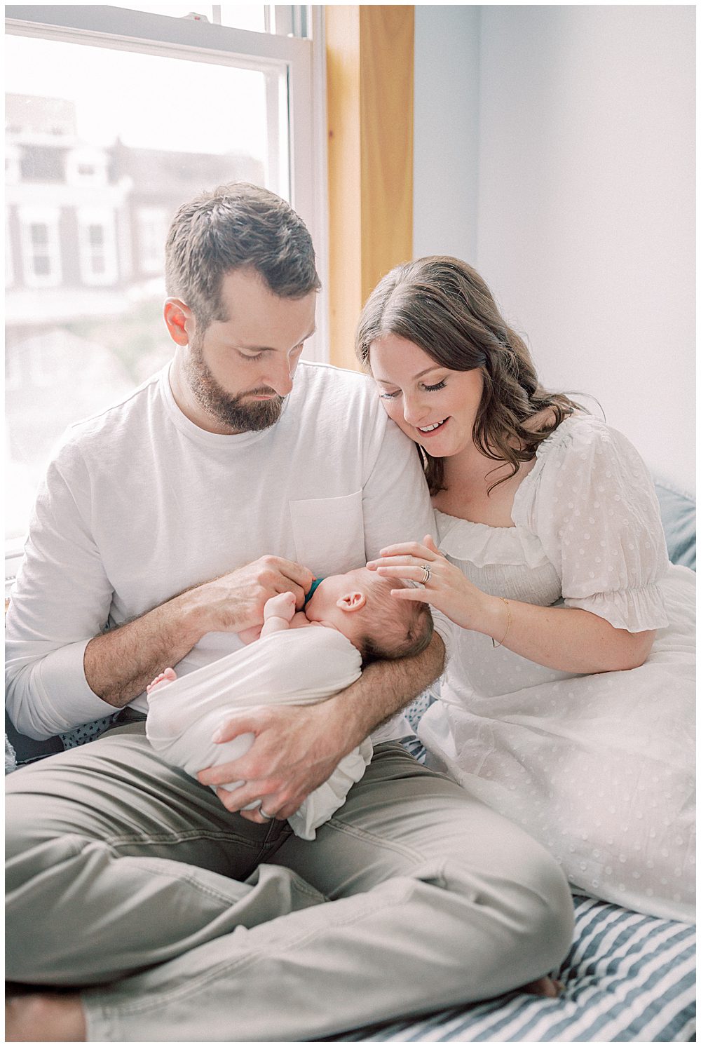 Dad Sits On Bed And Holds His Newborn Son While Wife Leans Against Him, Gently Rubbing Newborn's Head During Their Capital Hill Newborn Session.