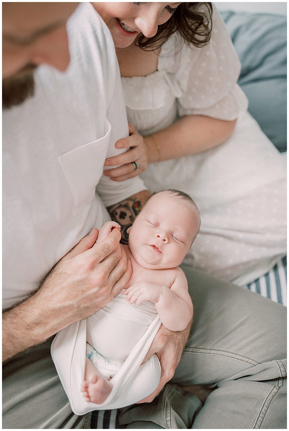 New Parents Hold Their Newborn Baby Boy In Their Lap While Sitting On Their Bed