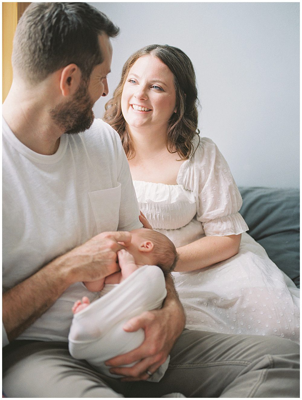 New Mom Smiles At Her Husband While He Holds Their New Baby While They Sit On Their Bed