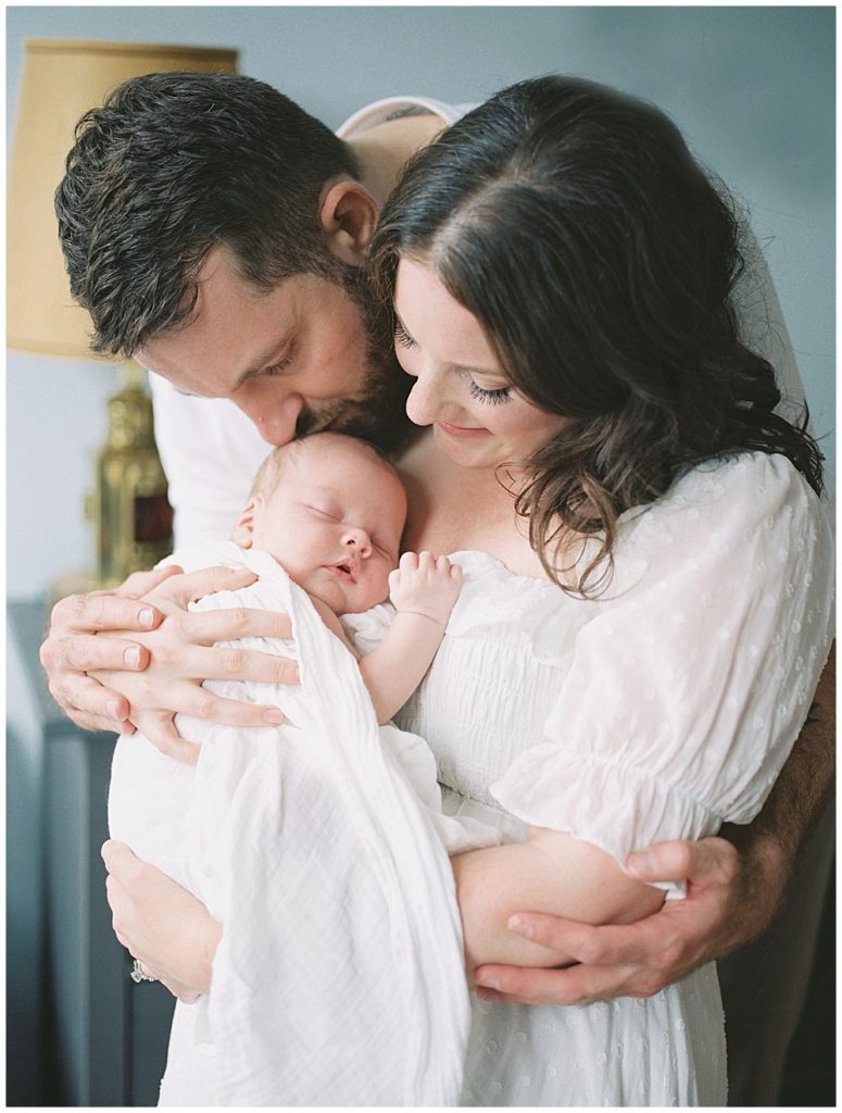 New Father Leans Over His Wife's Shoulder To Kiss His Newborn Baby.