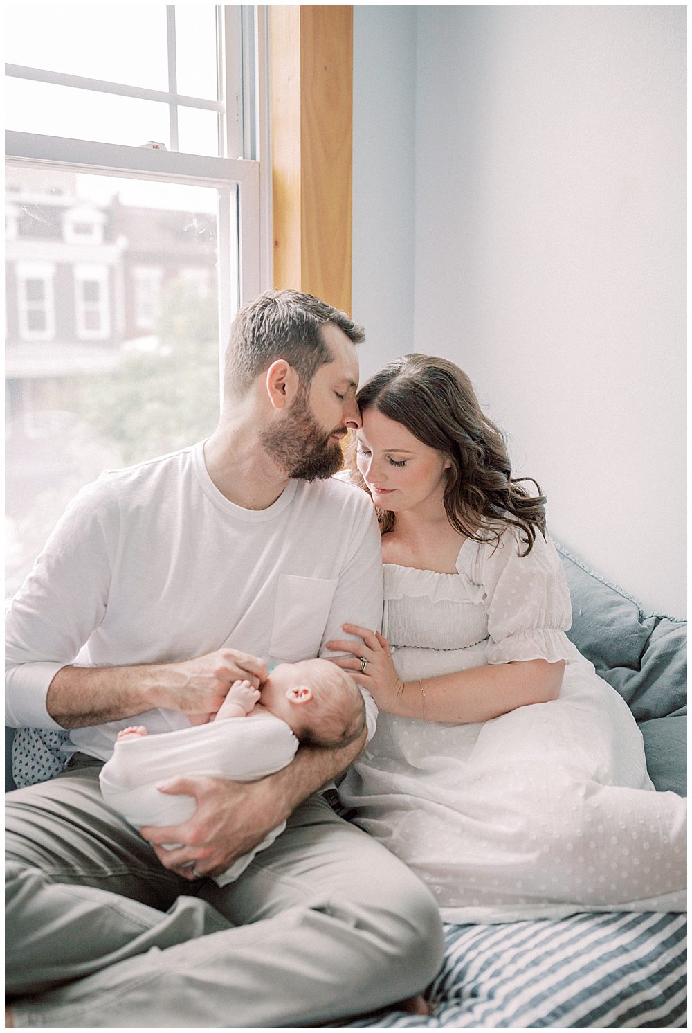 Father Kisses The Forehead Of His Wife While They Sit On Their Bed Holding Their Newborn Baby During Their Capital Hill Newborn Session