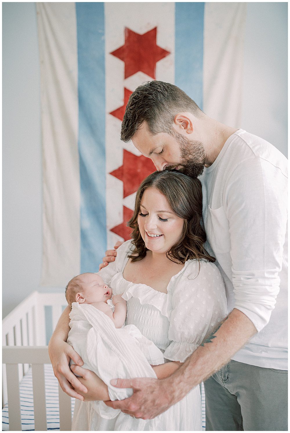 New Dad Leans Over His Wife And Kisses Her On The Head While She Holds Their Newborn Baby In Front Of Their Chicago-Themed Nursery