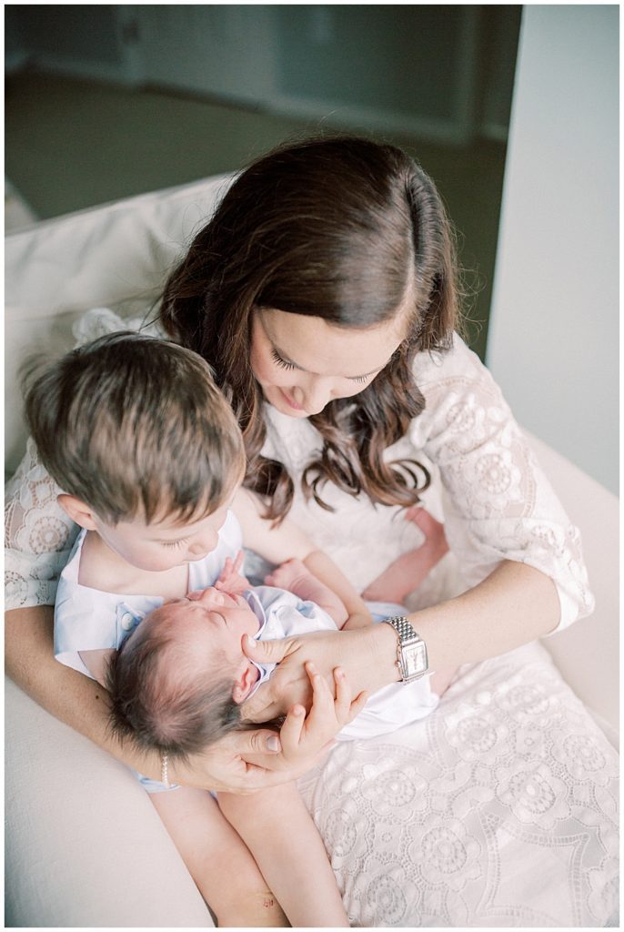 Mother Holds Her Toddler Son And Newborn Baby In Her Lap In An Ivory Chair.