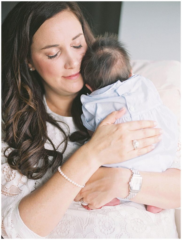 Mom Holds Up A Baby Boy In Blue Outfit And Fuzzy Hair Up To Her Cheek.