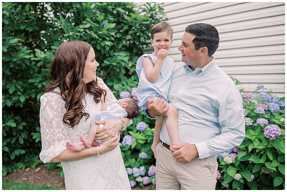 Little Boy Held By His Father Smiles While His Mother Holds His Baby Brother In Front Of The Hydrangea Bush.