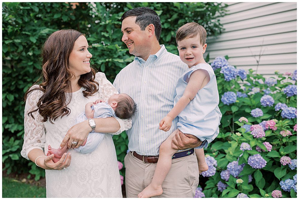 Toddler Boy Smiles At The Camera While Being Held By His Parents With Their Newborn Son.