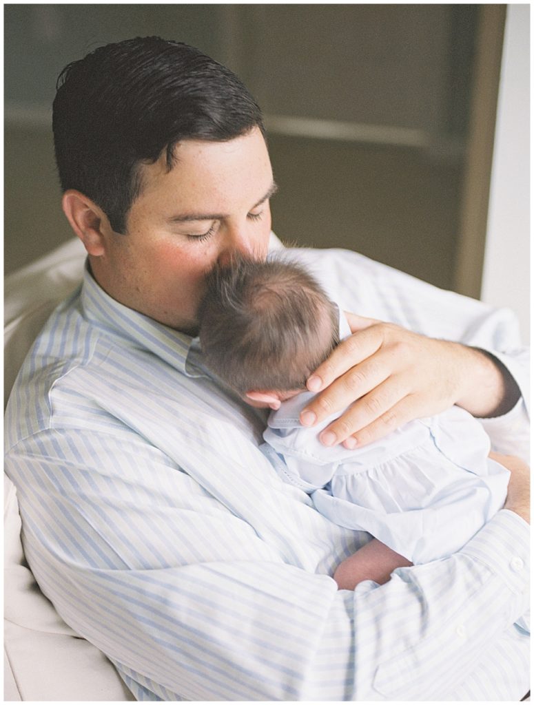 New Dad Brings His Newborn Son Up To Kiss Him On The Head While Sitting In A Chair.