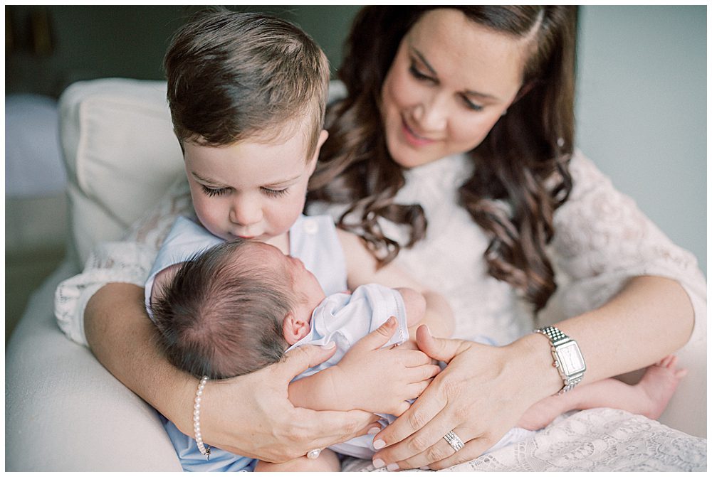 Toddler Boy Kisses His Baby Brother On The Head While Both Are Held By Their Mother.