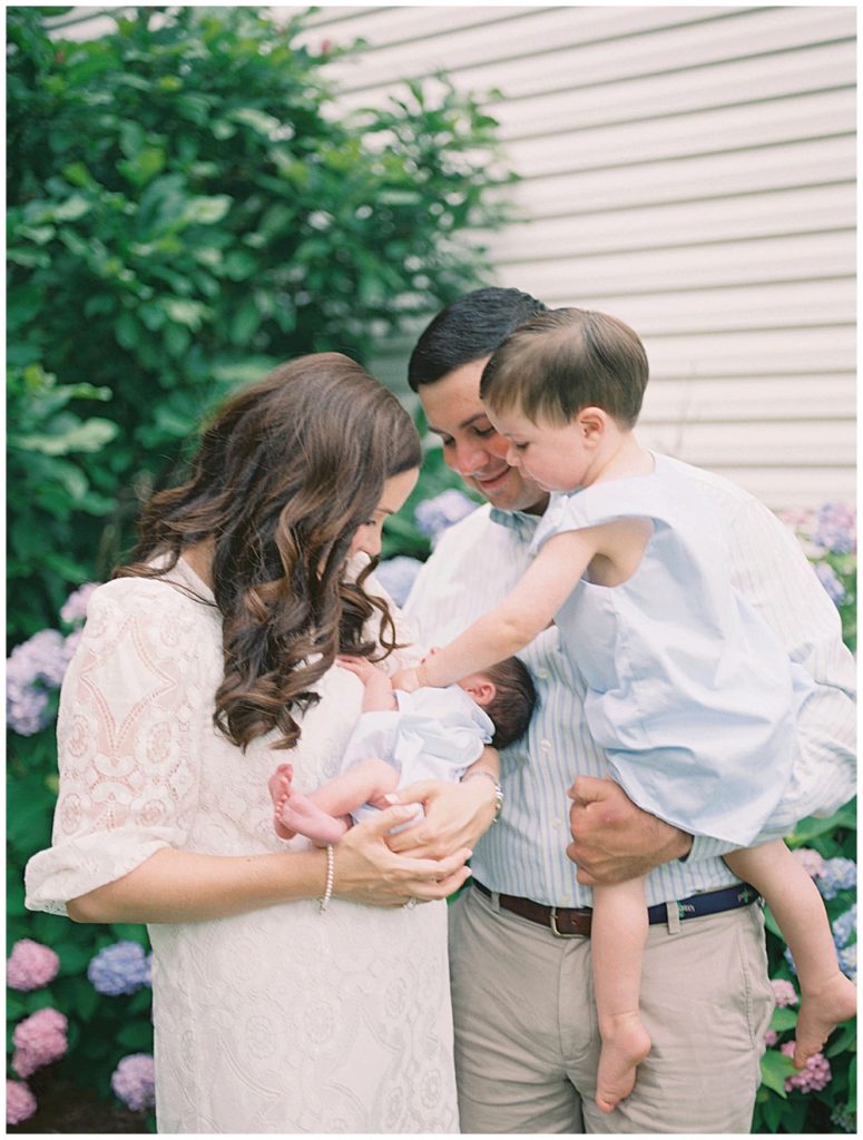 Toddler Boy Held By Dad Leans Over To Touch His Newborn Brother Held By His Mother During Their Fairfax Newborn Session.