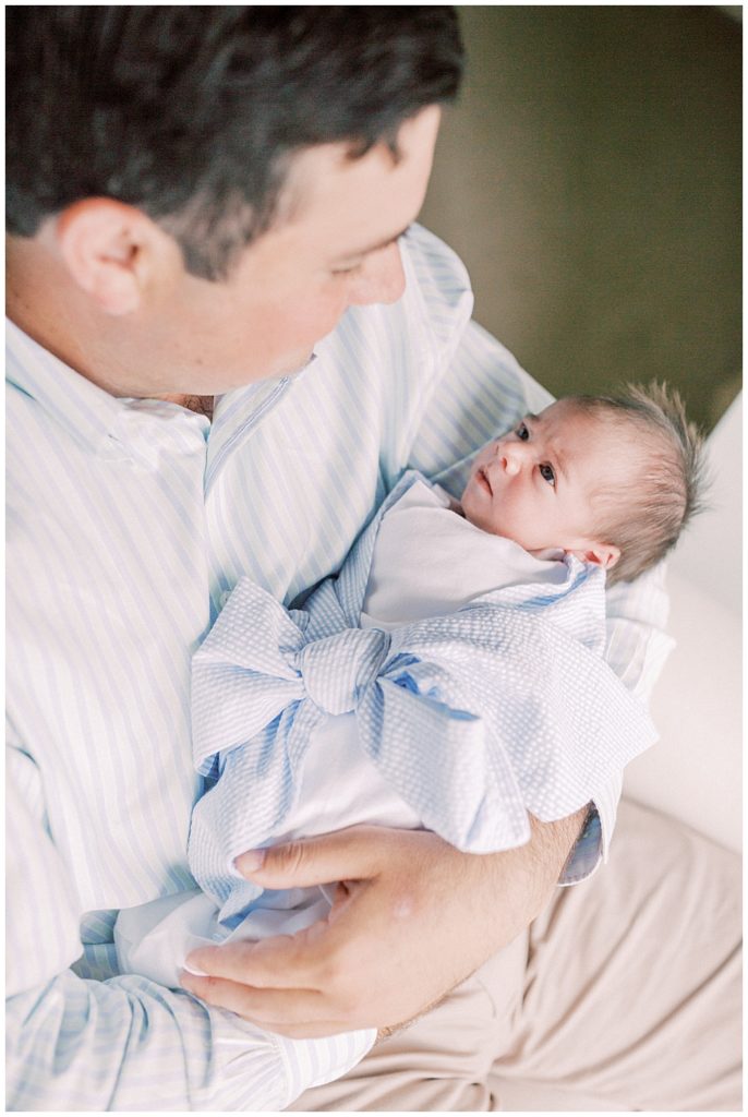 Newborn Baby Boy Wrapped In Beaufort Bonnet Company Blue Bow Swaddle Looks Up At His Father.