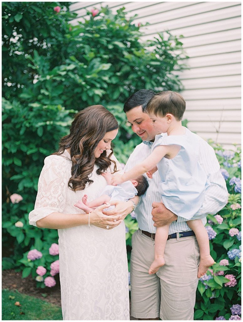 Toddler Boy Held By His Father Leans Over To Touch His Baby Brother Held By His Mother During Their Fairfax Newborn Session.