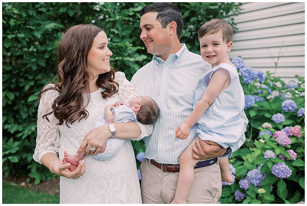 Mother And Father Smile At Each Other While Holding Their Toddler Boy And Newborn During Their Fairfax Newborn Session.