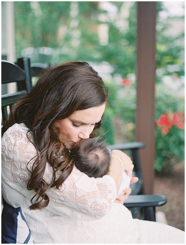 Brown-Haired Mom In White Dress Brings Her Newborn Baby Up For A Kiss On The Head While Sitting In A Rocking Chair Outside.