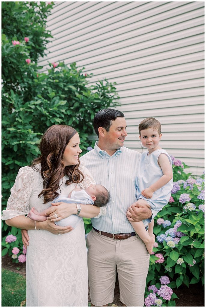 Mother And Father Smile At Their Toddler While Holding Both Their Toddler And Newborn Baby While Standing In Front Of Hydrangeas.