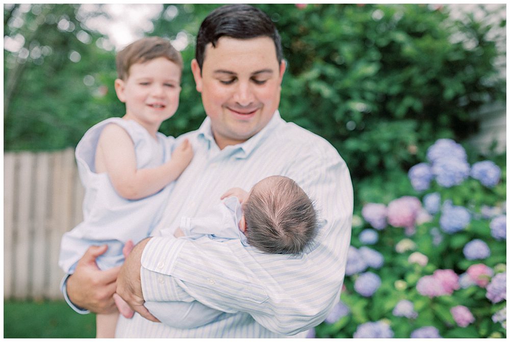 Father Smiles Down At His Newborn Baby While Holding His Newborn And Toddler.