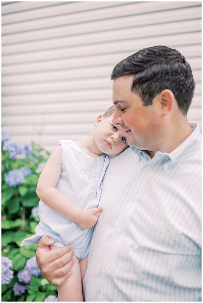 Toddler Boy Lays His Head Against His Father's Shoulder While They Stand In Front Of A Hydrangea Bush.