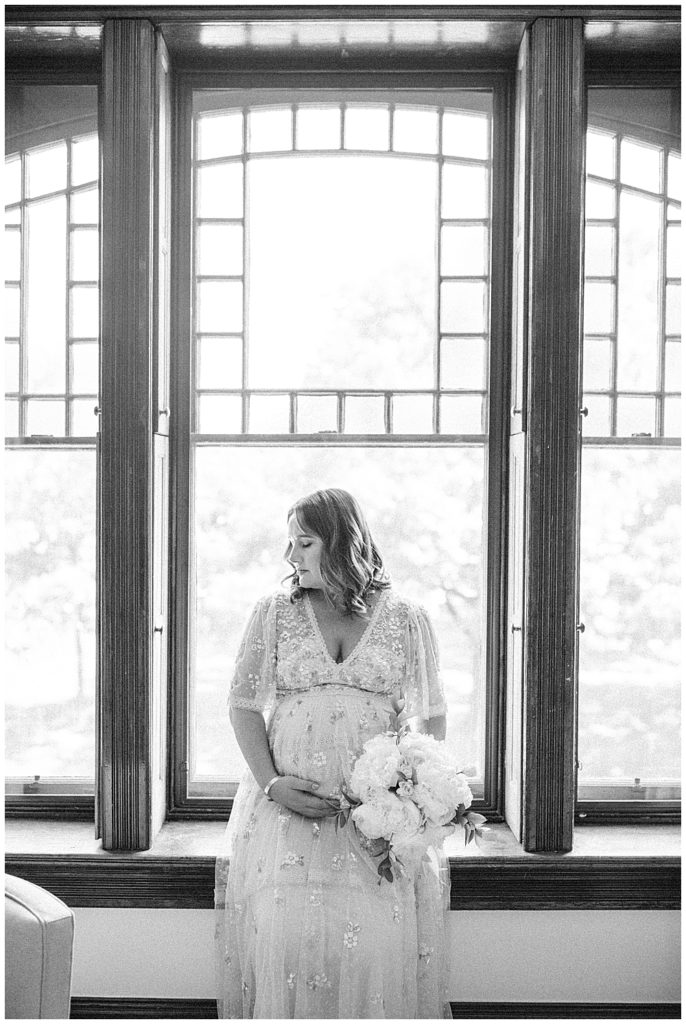 Black And White Image Of Pregnant Mom Sitting In Front Of A Large Transom Window Holding Both Her Belly And A Bouquet