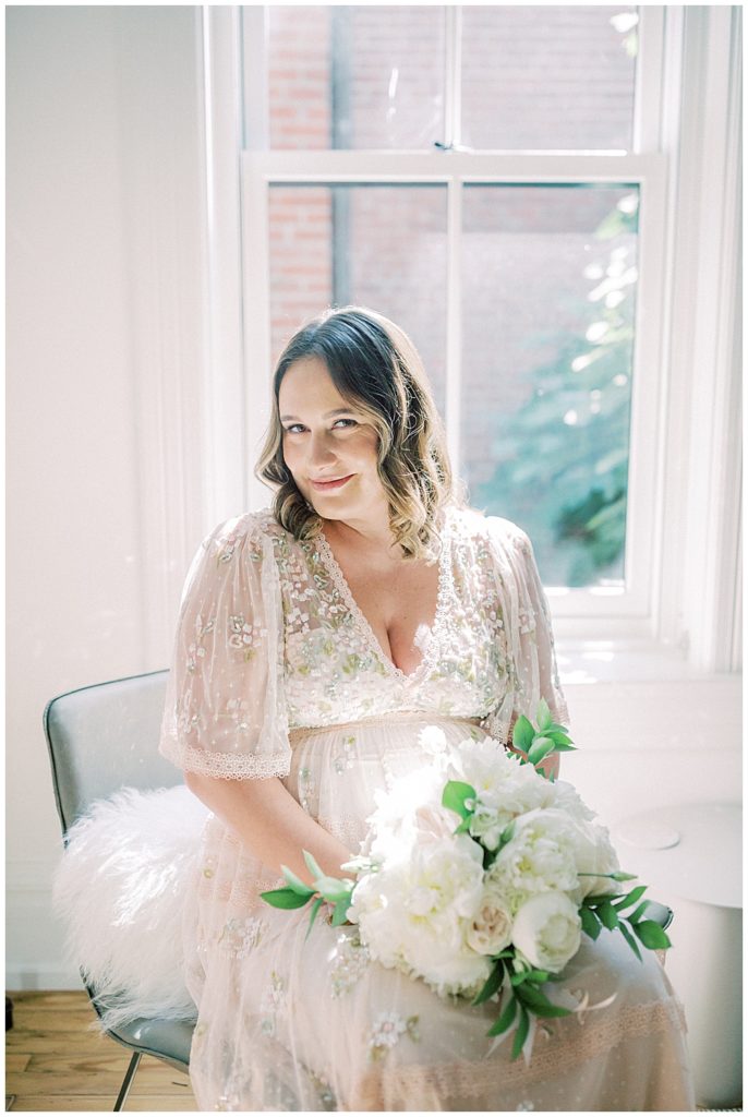 Pregnant Mom Sits In A Chair Wearing A Pink Needle &Amp; Thread Gown And Holds A Bouquet Of White Roses