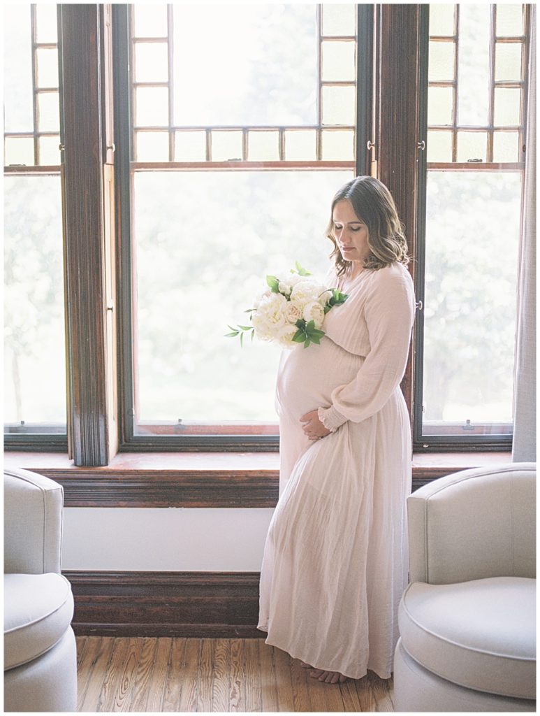 Mom Stands In Window Frame Holding Bouquet Of White Roses And Places One Hand Below Her Pregnant Belly