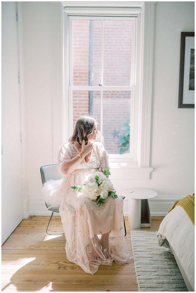 Pregnant Mom Sits In A Chair Wearing A Pink Needle &Amp; Thread Sequin Gown And Looks Out The Window While Holding A Bouquet Of White Roses And Brushing Hair From Her Face