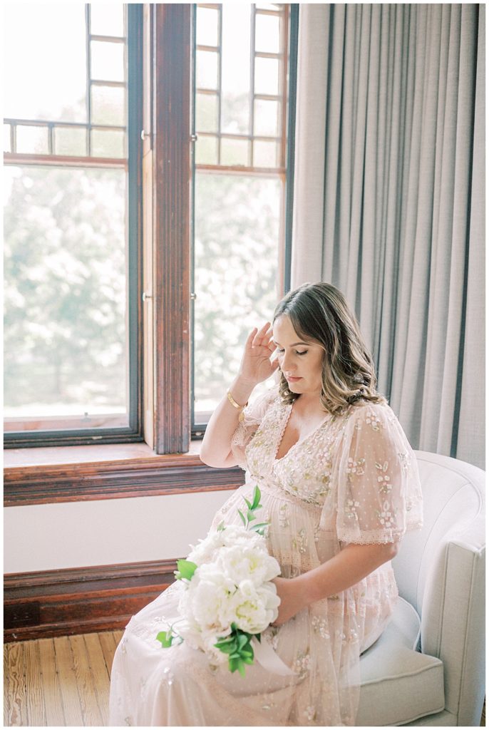 Expecting Mother Sits In A Chair Wearing A Pink Needle &Amp; Thread Gown And Holding A Bouquet Of White Roses While Brushing Hair Away From Her Face.