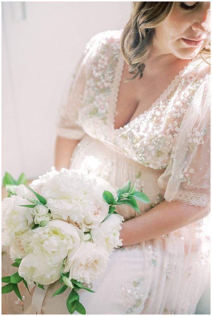 View Of Pregnant Mom In A Pink Needle And Thread Dress Sitting Holding A Bouquet Of White Flowers