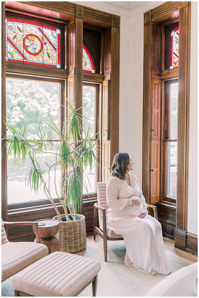 Pregnant Woman Sits In Chair In Her Home Looking Out The Window During Her In-Home Maternity Session