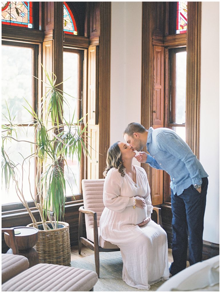 Pregnant Mom Sits In A Chair Holding Her Belly While Her Husband Leans Down To Kiss Her