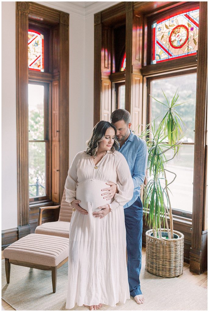 Expecting Mother Stands In Front Of Her Husband While They Both Have Their Hands On The Belly Of The Mother