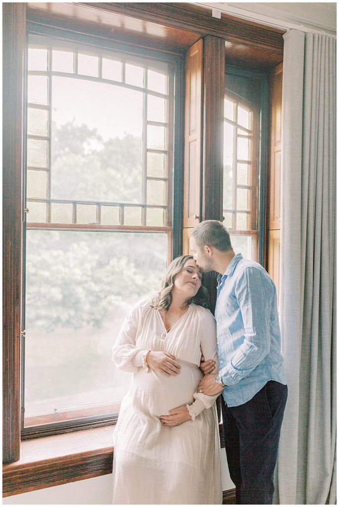 Husband Leans Over To Kiss Pregnant Wife's Forehead While She Sits In The Window Ledge Of Their Home