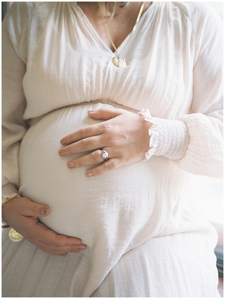 Pregnant Mom Wearing An Ivory Dress Places Hand On Her Belly