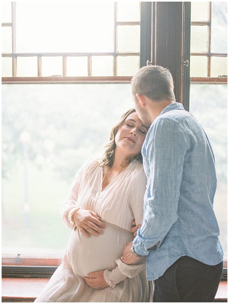 Expecting Mother Sits In Her Windowsill While Her Husband Gently Kisses Her Forehead During An In-Home Maternity Session In Washington Dc