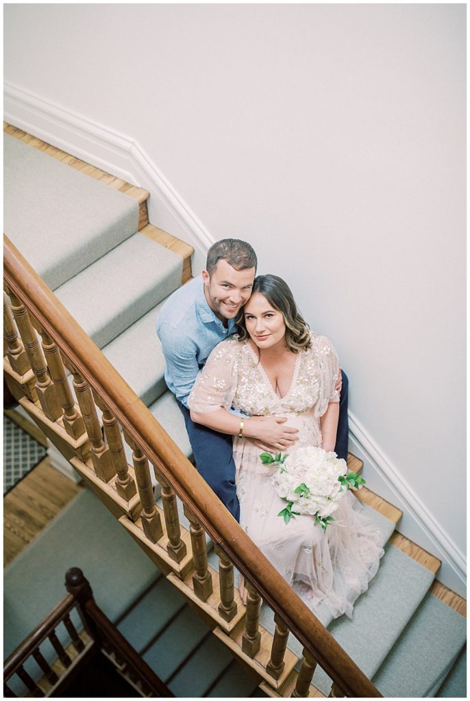 In-Home Maternity Session Where Expecting Mother And Her Husband Sit On Their Steps Looking Up At The Camera