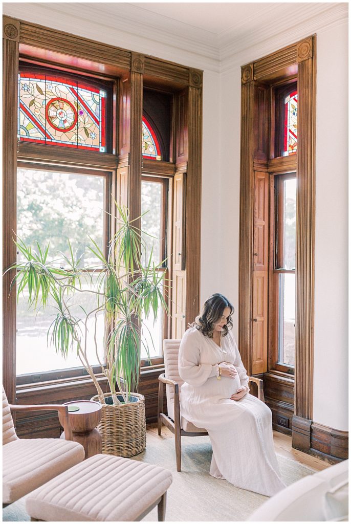 Pregnant Mother Sits In A Chair In Her Home Looking Down At Her Belly Surrounded By Tall Windows With Red Transom Windows And Wooden Paneling 