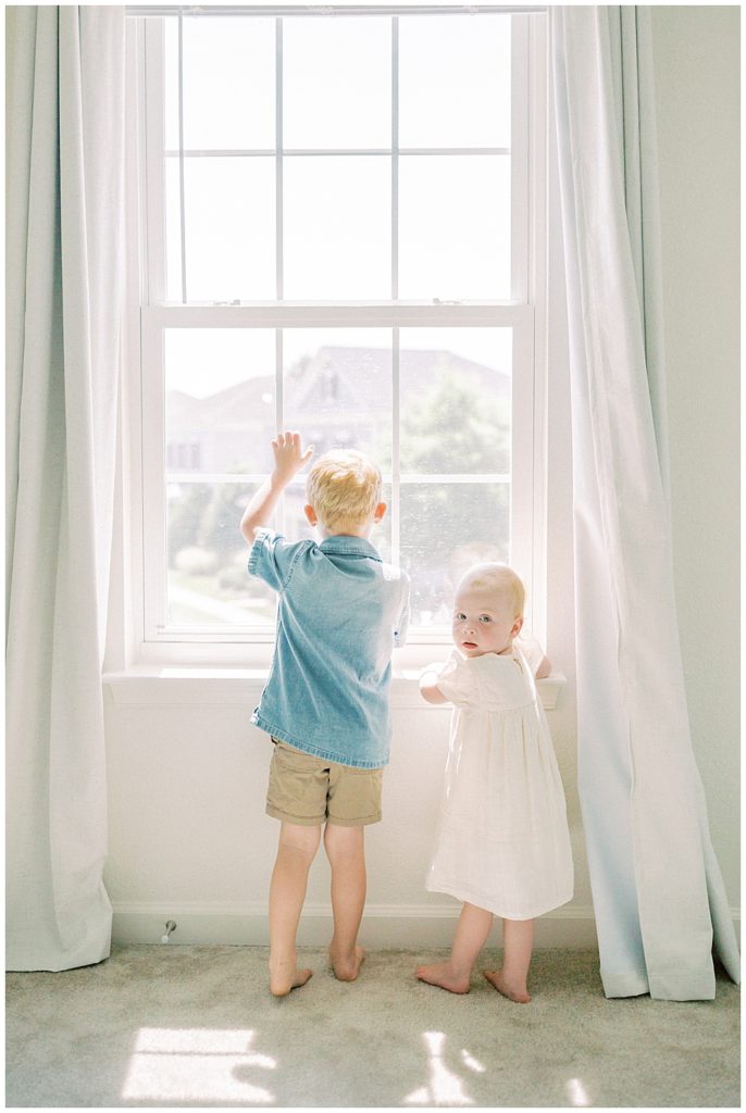 Toddler Girl Stands With Her Bigger Brother In The Window And Looks Back At The Camera.